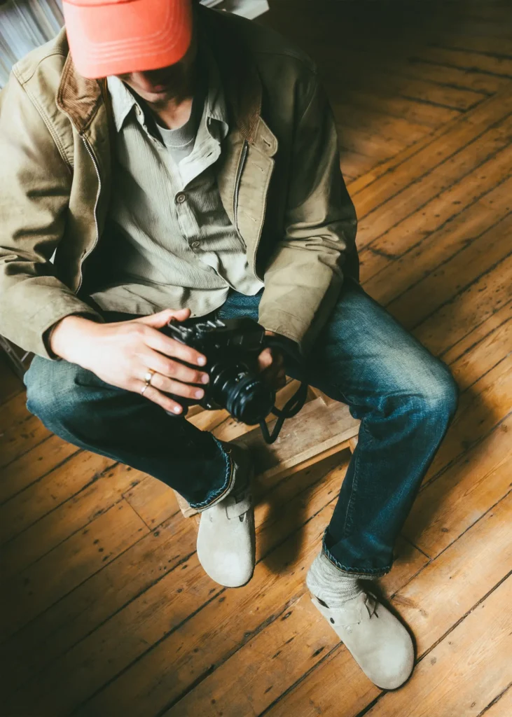 Overhead lifestyle image of a person seated on a low stool in a warm, loft-style interior, wearing light beige suede clogs with grey socks, relaxed denim, layered neutral workwear, and a red cap while holding a camera. Natural light and worn wooden flooring create a calm, lived-in atmosphere that emphasizes creativity, comfort, and everyday utility style