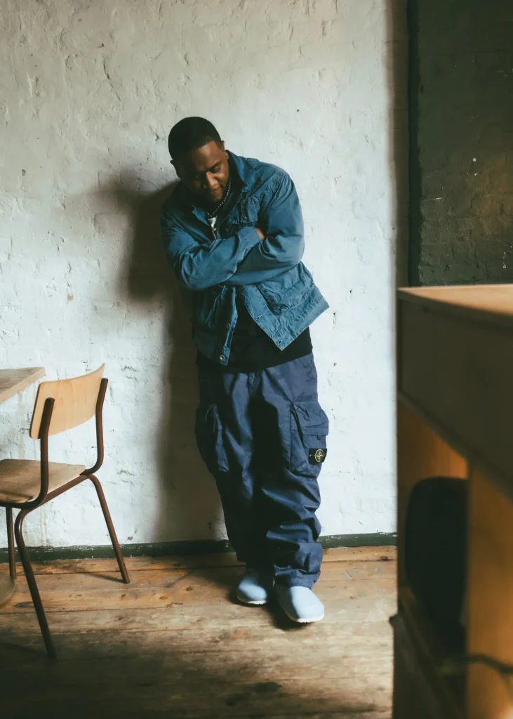 Lifestyle image of a person standing casually against a textured plaster wall in a warm industrial-style interior, wearing light blue slip-on clogs with relaxed cargo trousers and layered blue outerwear. The scene combines soft natural light, worn wooden flooring, and understated styling to emphasize comfort, utility, and day wear