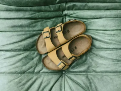 Overhead lifestyle image of a pair of mustard-yellow two-strap sandals arranged on a softly quilted sage-green textile surface. The composition highlights the contrast between textured leather, worn cork footbeds, and plush fabric, creating a calm, tactile editorial scene centered on comfort and understated design