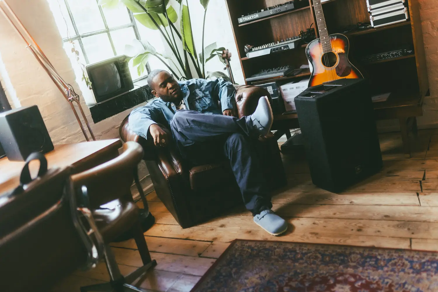 Lifestyle image of a person relaxing in a vintage-inspired creative studio, seated in a brown leather armchair while wearing light blue slip-on clogs, relaxed utility trousers, and layered blue outerwear. Surrounded by speakers, a guitar, music equipment, indoor plants, and warm wooden flooring, the scene creates an editorial atmosphere centered on comfort, music culture, and lived-in everyday style