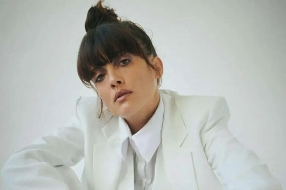 : Noga Erez poses in an oversized white suit and crisp button-up shirt against a minimal studio backdrop, leaning forward with a calm but piercing expression that reflects her sleek, avant-garde pop aesthetic