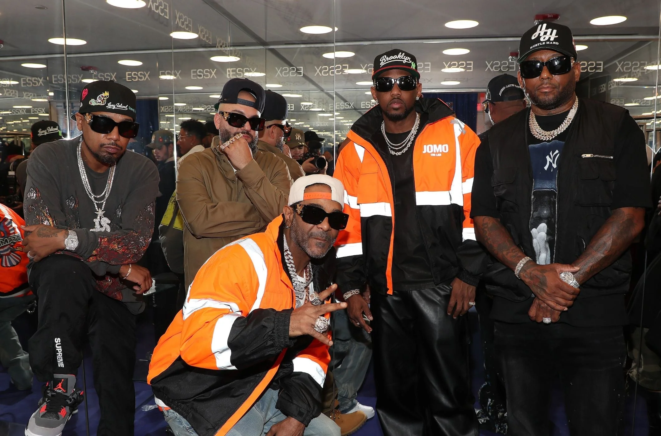 Members of a hip-hop collective pose together at the ESSX x Diplomats Vintage Frames launch party, wearing oversized sunglasses, layered diamond jewelry, baseball caps, and streetwear-inspired outfits. Two men in bright orange reflective jackets stand out against the mirrored interior, while the group gathers confidently for a fashion-forward portrait celebrating New York hip-hop culture and luxury eyewear