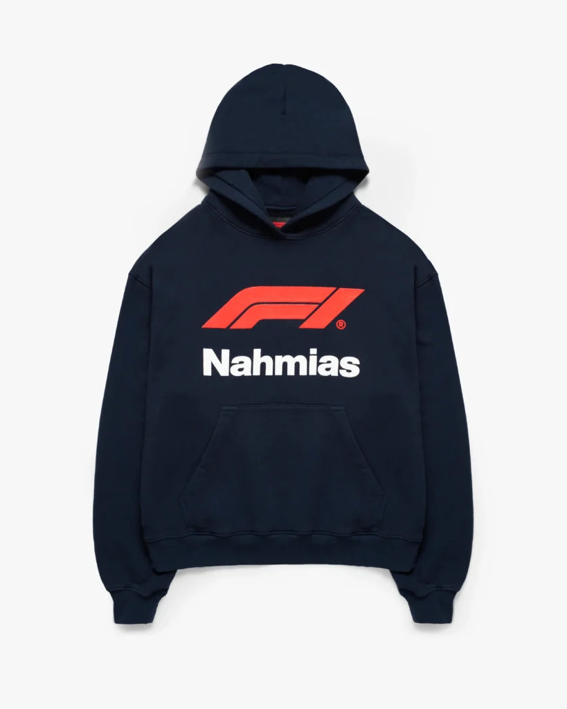 Navy pullover hoodie with a structured, slightly relaxed fit and a classic kangaroo pocket. The front is dominated by an oversized red Formula 1 logo, set above bold white “Nahmias” lettering, creating a high-contrast focal point. A spacious hood and ribbed cuffs and hem complete the silhouette, while the deep navy base grounds the vivid branding. The result is a clean yet statement-driven piece that merges motorsport iconography with elevated streetwear sensibility