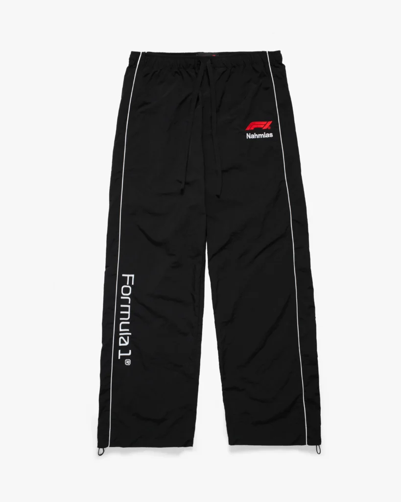 Black lightweight track pants with a relaxed, straight-leg silhouette and a slightly crinkled finish. The elastic waistband features an adjustable drawstring for a customizable fit, while subtle white piping runs down each leg, adding a clean, streamlined contrast. The right thigh is marked with a red Formula 1 logo above small “Nahmias” branding, while the left leg features bold vertical “Formula 1®” text. Adjustable cords at the hems allow for a tapered or loose fit, completing the motorsport-inspired design with functional, street-ready versatility