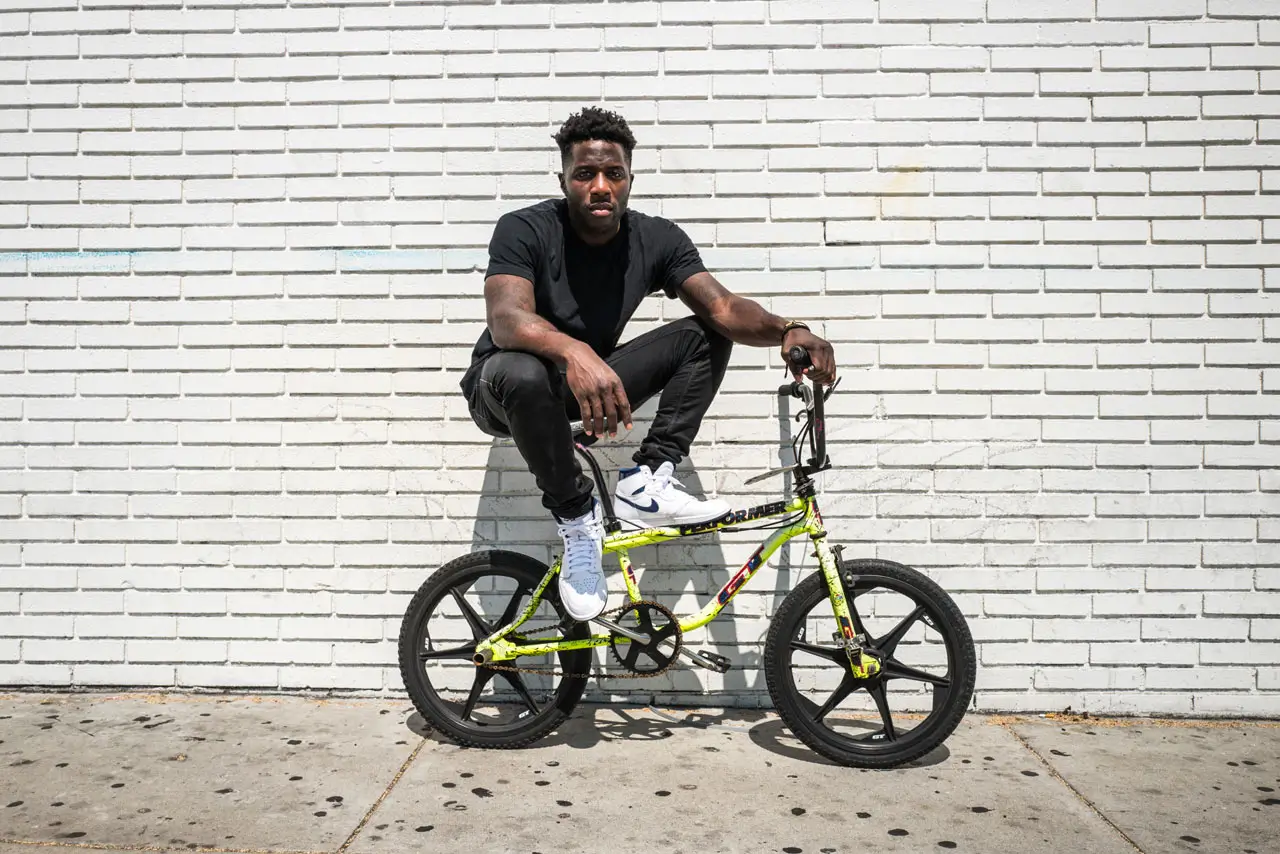 An outdoor portrait of Nigel Sylvester seated casually on a neon yellow BMX bike in front of a long white-painted brick wall under bright daylight. Dressed in an all-black outfit with white Nike sneakers, Sylvester balances across the bike frame with a relaxed but focused posture, reflecting the understated confidence associated with his BMX and streetwear identity. The weathered sidewalk, stark brick backdrop, and vivid bike graphics create a raw urban atmosphere that mirrors the gritty, progression-driven ethos behind his “Bike Air” collaborations and wider creative universe