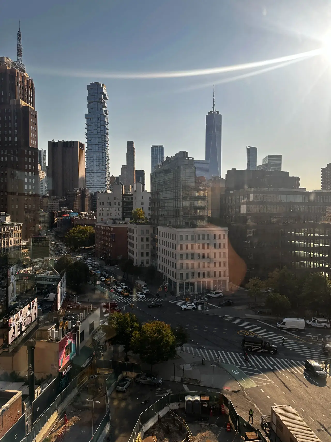 Sunlit New York City skyline viewed from above a busy intersection, with One World Trade Center rising in the distance, modern glass towers and mid-rise buildings layered across the scene, long shadows cast by late-afternoon light, and traffic flowing through crosswalk-lined streets below