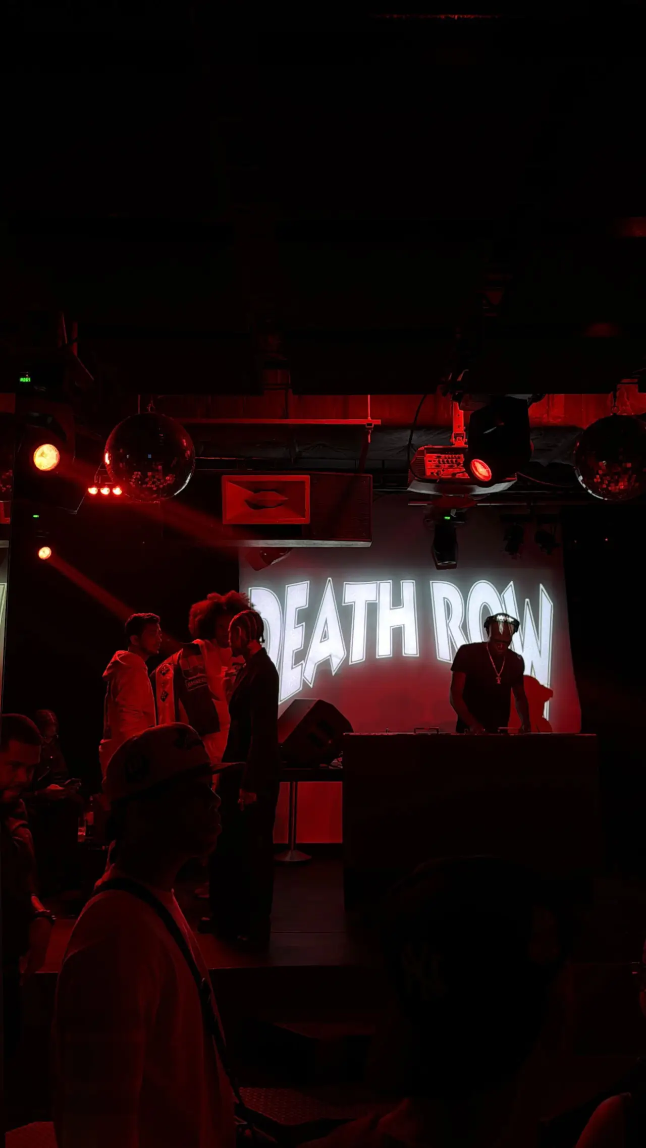 Dimly lit nightclub scene washed in deep red light, with a DJ performing behind a booth beneath a glowing “DEATH ROW” backdrop, while small groups of people stand and talk on stage and in the crowd, mirrored disco balls and stage lights hanging overhead, creating a moody, intimate live music atmosphere
