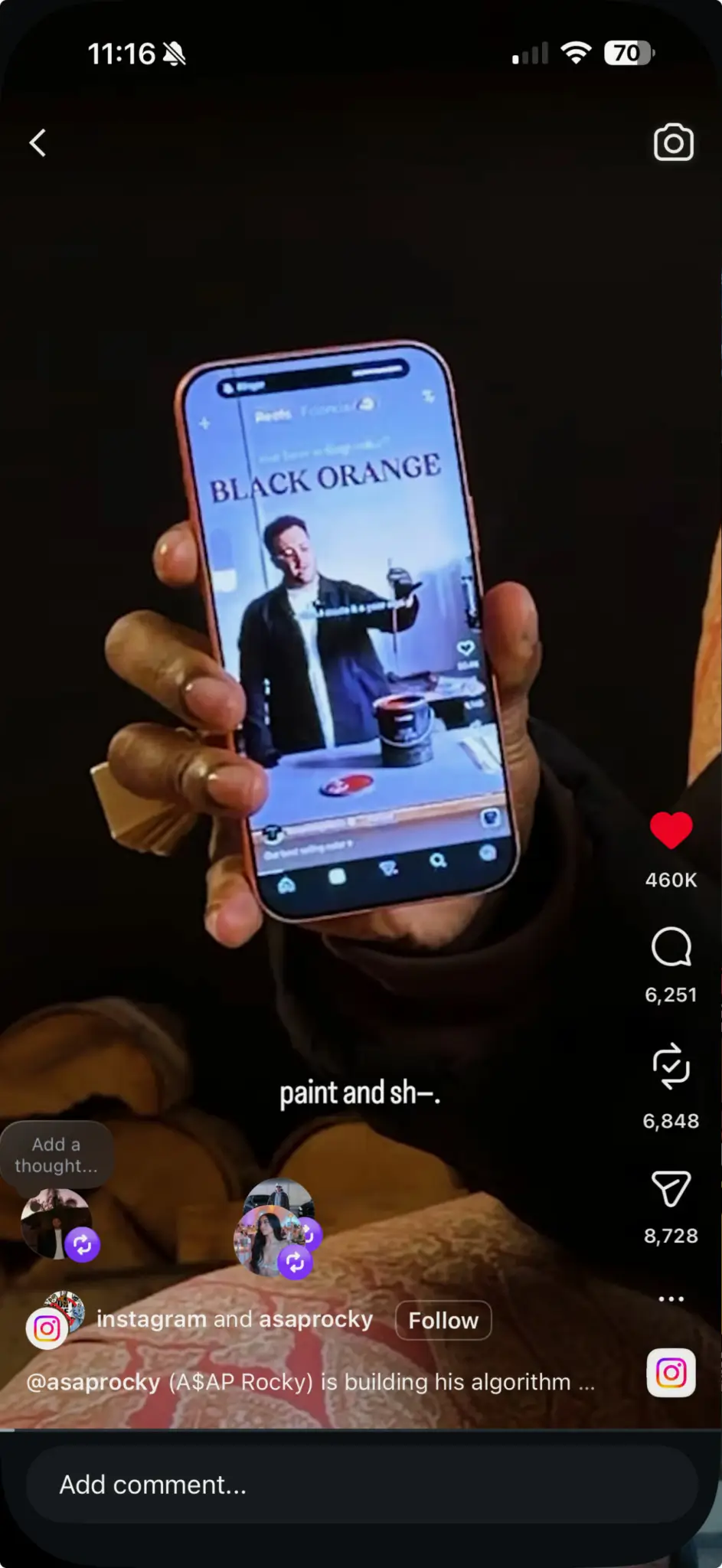 Close-up of a person holding a smartphone displaying a social media video, where a man demonstrates paint labeled “BLACK ORANGE,” with on-screen interface elements showing likes, comments, and share icons, while the viewer’s hand and part of their clothing are visible in a dim indoor setting