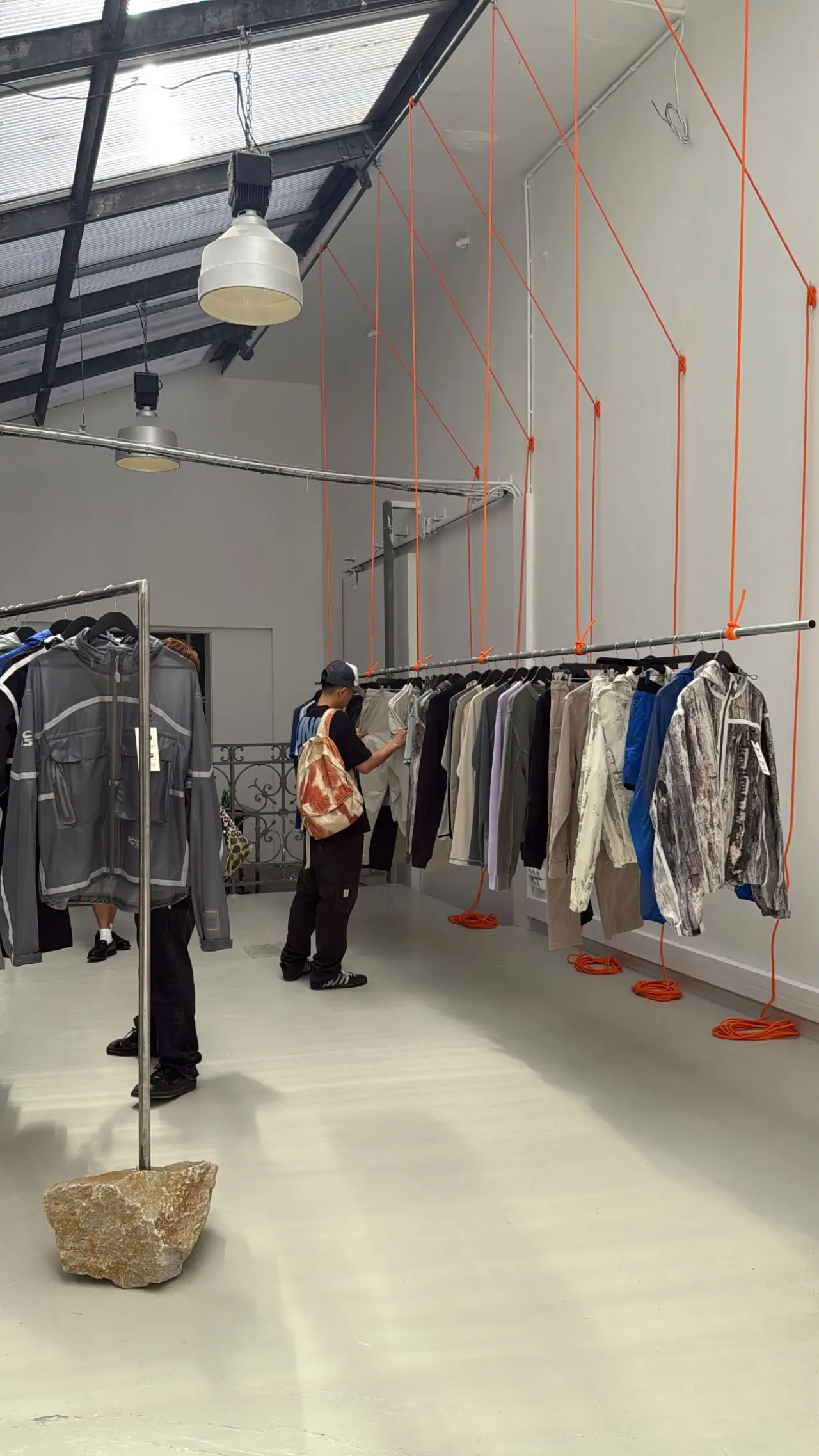 Minimalist fashion showroom with a high industrial ceiling and skylights, where clothing racks are suspended from the ceiling by bright orange cords, displaying a curated selection of jackets and garments. A person wearing a cap and backpack browses the pieces, while another rack stands nearby, anchored by a large stone base, reinforcing the space’s clean, conceptual retail design