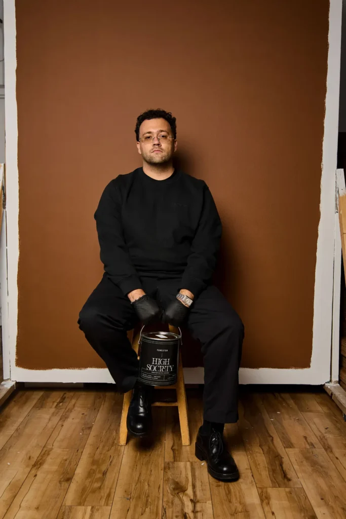 Portrait of a man seated on a wooden stool in a studio, dressed in all black with glasses and gloves, positioned in front of a freshly painted brown wall with visible edges. A paint can labeled “HIGH SOCIETY” rests between his knees, set on a wooden floor, giving the scene a composed, minimalist, and work-in-progress aesthetic