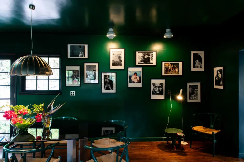 Moody interior dining space with deep green walls, featuring a gallery arrangement of framed photographs and artwork, softly lit by pendant and wall-mounted lights. A glass dining table with chairs sits beneath a sculptural hanging lamp, accented by a vase of bright flowers, while wooden floors and a nearby seating corner add warmth and a curated, lived-in atmosphere