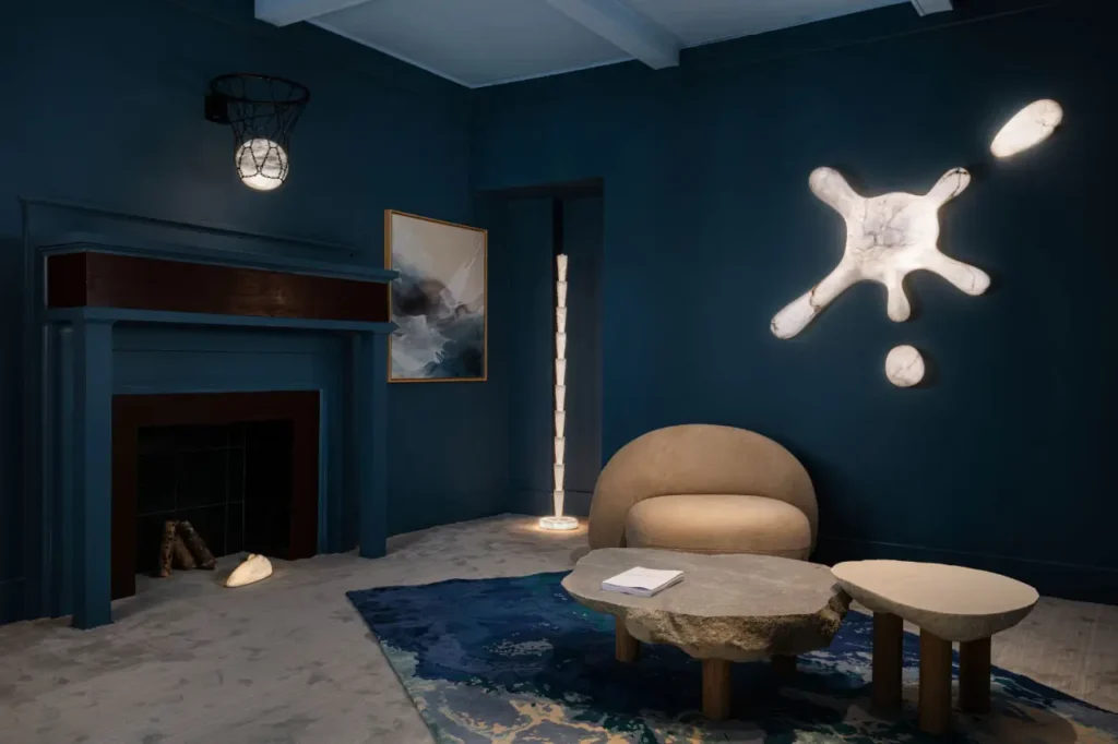 Stylish, moody living room with deep blue walls, featuring a sculptural wall light resembling an abstract splash, a rounded upholstered lounge chair, and organic stone tables set on a textured rug. A modern floor lamp and framed artwork complement the space, while a minimalist fireplace and soft ambient lighting create a refined, gallery-like interior atmosphere