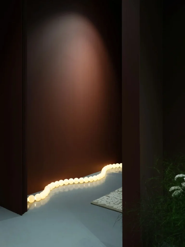 Moody, minimalist interior scene featuring deep brown walls and a soft spotlight glow, with a winding line of small illuminated bulbs tracing along the floor, creating a sculptural light installation. A partial view of greenery and white flowers appears at the edge, adding contrast to the otherwise controlled, design-forward composition