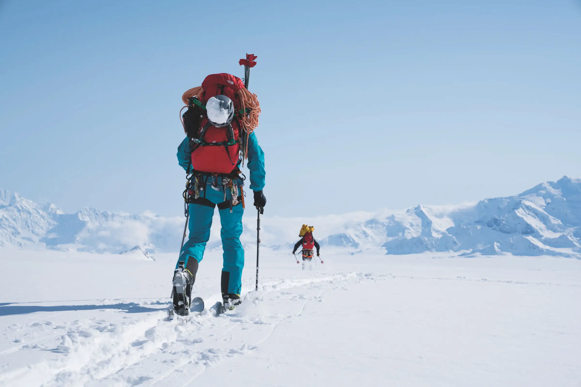 Expedition climbers traverse a vast snow-covered glacier carrying heavy mountaineering packs and technical gear beneath expansive alpine skies