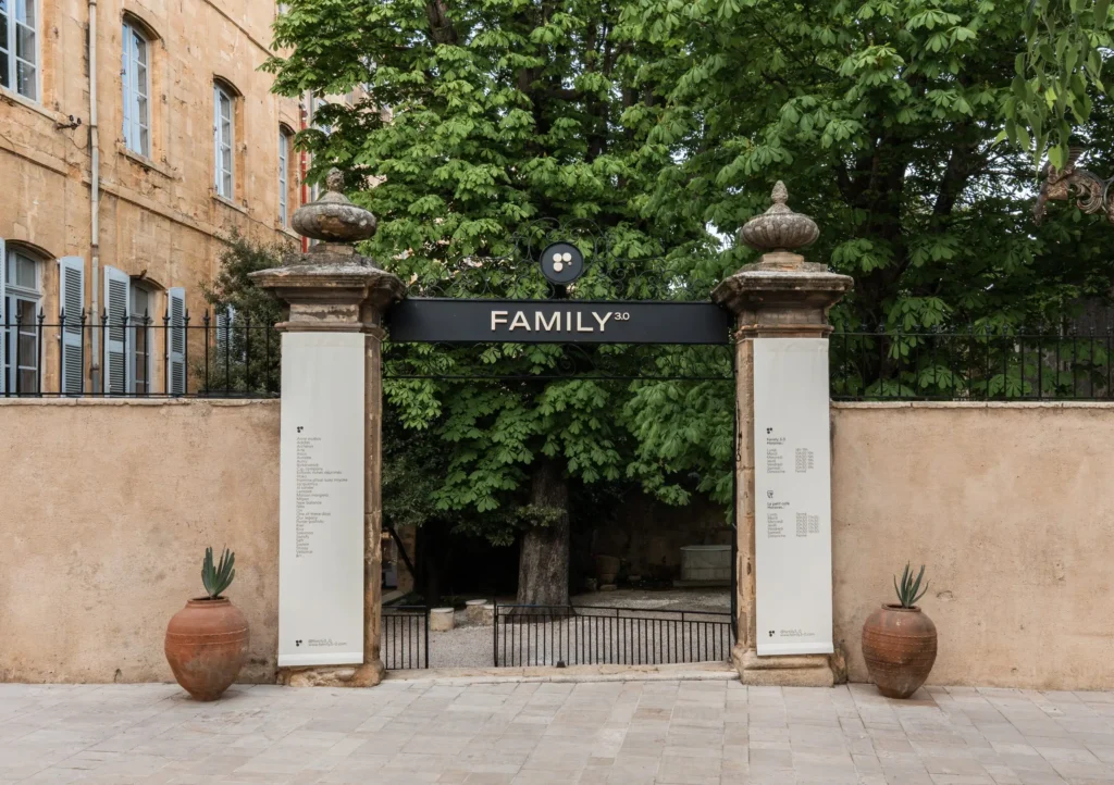 Full exterior entrance to Family 3.0 featuring a classic stone gateway with ornate wrought iron detailing, central branded signage, flanked by sculptural columns and greenery leading into a courtyard setting