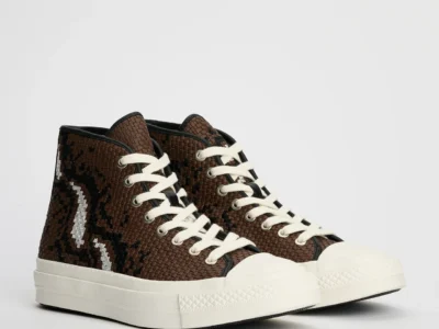 Pair of brown Converse First String Chuck Taylor high-top shoe displayed against a clean light grey studio background, featuring intricately woven leather-style uppers with black and metallic silver abstract patterning, thick off-white rope laces, black piping details, and chunky off-white vulcanized rubber soles that emphasize the silhouette’s elevated craft-focused construction
