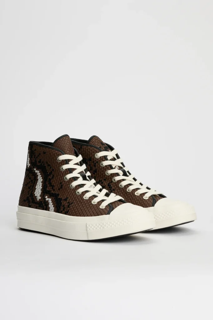 Pair of brown Converse First String Chuck Taylor high-top shoe displayed against a clean light grey studio background, featuring intricately woven leather-style uppers with black and metallic silver abstract patterning, thick off-white rope laces, black piping details, and chunky off-white vulcanized rubber soles that emphasize the silhouette’s elevated craft-focused construction