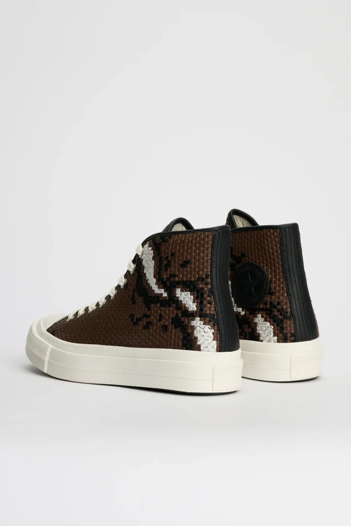 Rear angled view of a pair of Converse First String Chuck Taylor high-top shoe in brown, featuring tightly woven leather-style uppers with black and metallic silver abstract detailing, black textured heel panels, signature Converse ankle patch branding, off-white rope laces, and clean cream rubber soles set against a minimal light grey studio backdrop