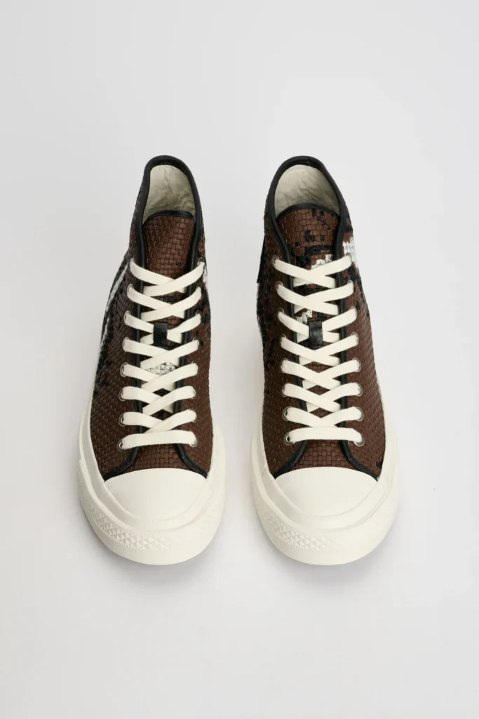 Top-down studio view of a pair of Converse First String Chuck Taylor high-top shoes in brown, showcasing tightly woven leather-style uppers with subtle black and metallic silver detailing, thick off-white rope laces, black trim accents, and clean cream rubber toe caps and soles against a minimal light grey background
