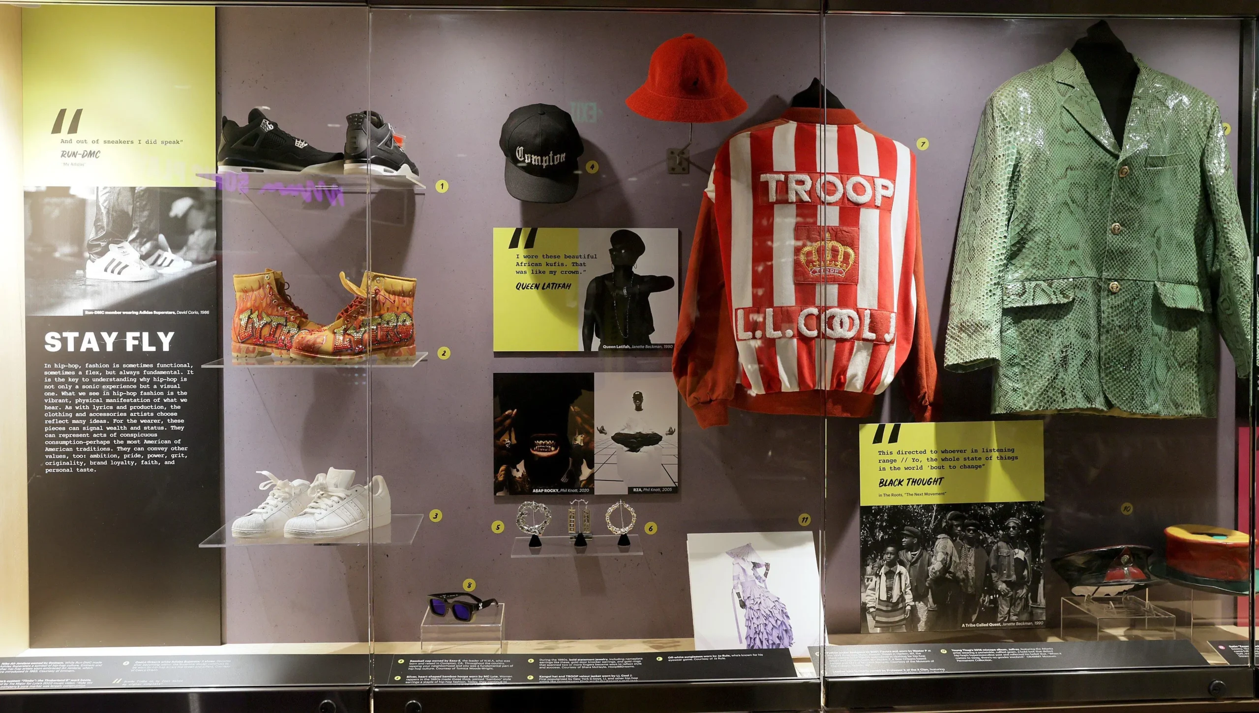 Archival-style fashion display inside the GRAMMY Museum showcasing hip-hop cultural memorabilia, including vintage sneakers, embellished boots, oversized jewelry, a red-and-white LL Cool J Troop jacket, Compton cap, and statement performance garments tracing the visual evolution of rap fashion and streetwear identity