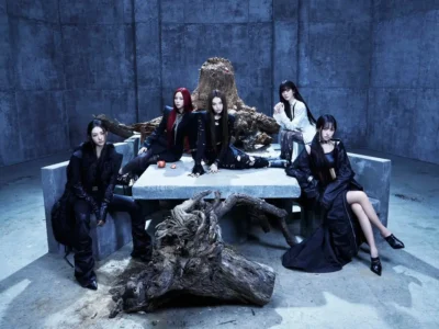 MEOVV poses in a dark industrial chamber for the BITE NOW promotional campaign, seated around a concrete table surrounded by twisted tree roots and fallen wood. The members wear predominantly black fashion-forward outfits with gothic-inspired styling, while cool blue lighting and stark concrete walls create a cinematic atmosphere. The composition blends themes of nature, decay, and mystery, reinforcing the EP’s fierce feline concept and dramatic view id