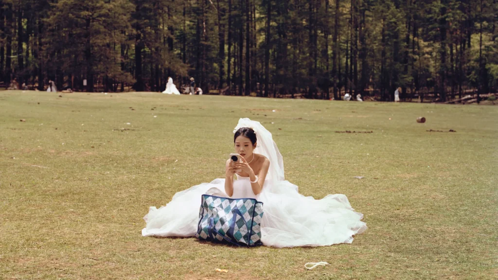 A bride in a voluminous white wedding gown sits alone in an open meadow checking her phone, with dense forest trees and distant wedding figures creating a quiet, cinematic scene of modern bridal tourism