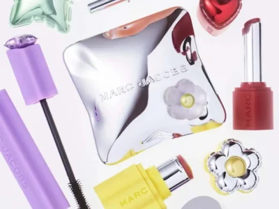 Marc Jacobs Beauty products displayed in a vibrant flat-lay composition, featuring chrome-finish compacts, a lavender mascara, pink eyeliner, yellow lipstick, and heart- and flower-shaped packaging. The playful mix of pastel colors, reflective metallic surfaces, and whimsical designs highlights the brand’s joyful new aesthetic for its 2026 beauty relaunch