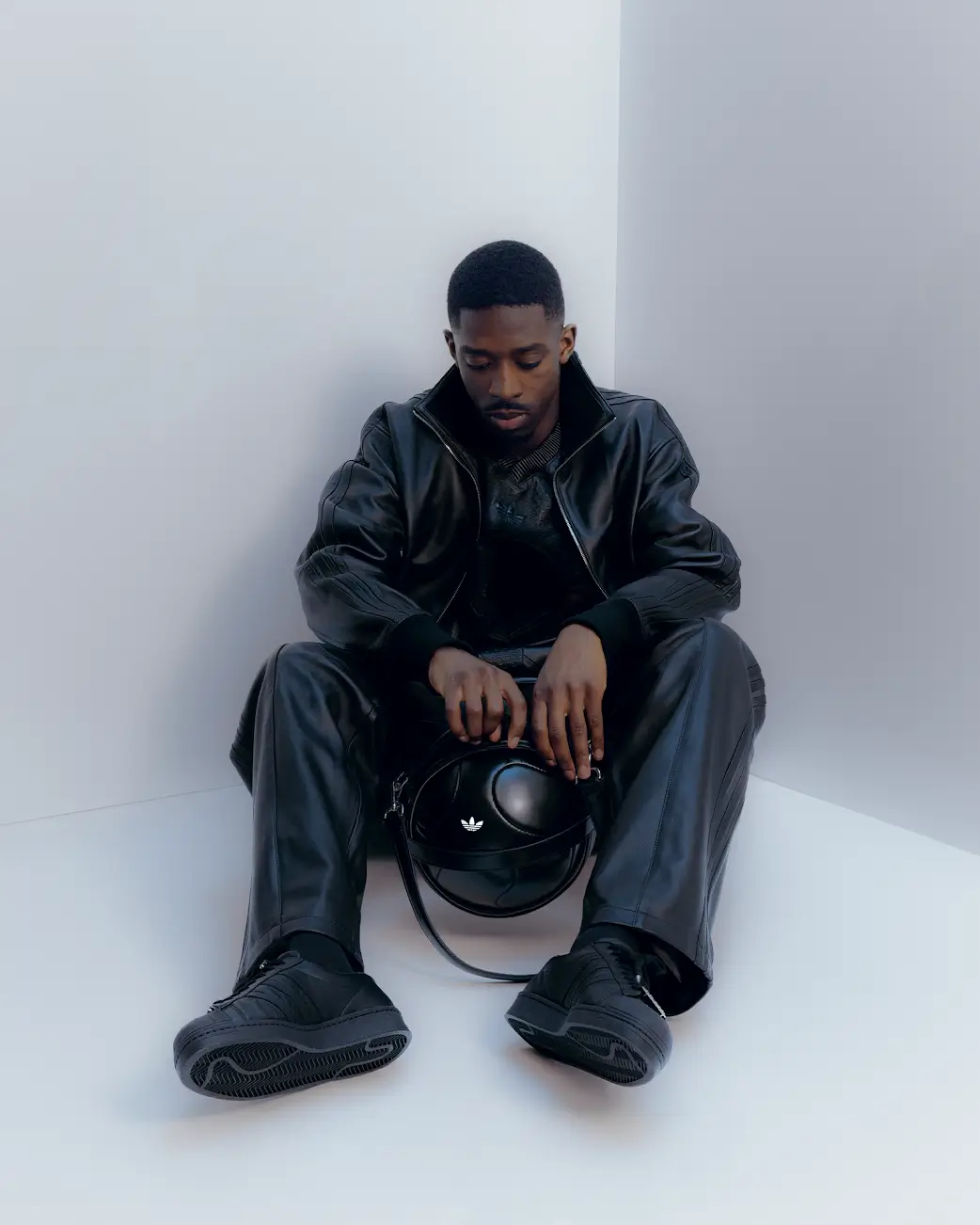 A fashion model sits inside a stark minimalist white studio corner wearing an all-black Adidas luxury sportswear ensemble composed of a leather track jacket, relaxed leather trousers, and monochromatic sneakers. Holding a sculptural black leather bag with subtle Trefoil branding, the look blends technical athletic heritage with elevated tailoring, while the soft cool-toned lighting and introspective pose create a sleek, futuristic editorial atmosphere