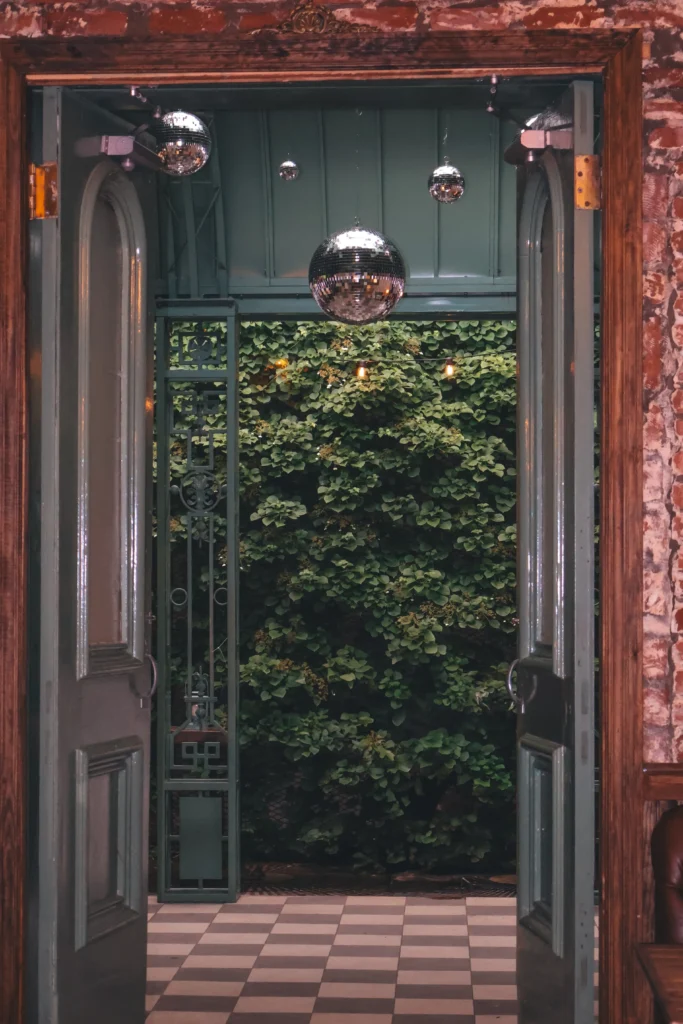 View through open vintage double doors into a lush garden courtyard, framed by exposed brick walls and checkerboard flooring, with mirrored disco balls suspended overhead creating a moody, romantic atmosphere inside Milk and Roses