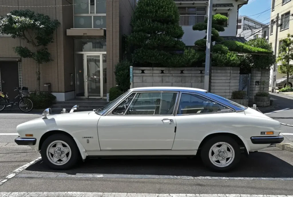 Side profile of a white Isuzu 117 Coupé showcasing its sleek fastback roofline, long hood, pillarless coupe design, and classic Japanese grand touring silhouette