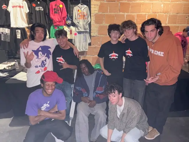Group photo of WHATMORE and friends gathered beside a merch booth, wearing casual streetwear and graphic tees while posing backstage in a DIY concert setting filled with hoodies, shirts, and underground tour energy