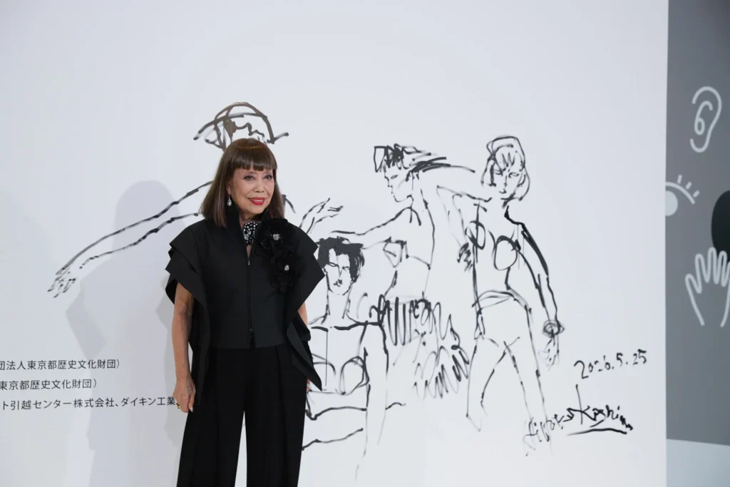 Portrait of a woman standing at the (UN)KNOWN HIROKO KOSHINO exhibition opening in front of a large white display wall featuring expressive black ink fashion sketches, wearing an architectural black ensemble with sculptural layered detailing that reflects the exhibition’s fusion of fashion, art, and personal creative expression
