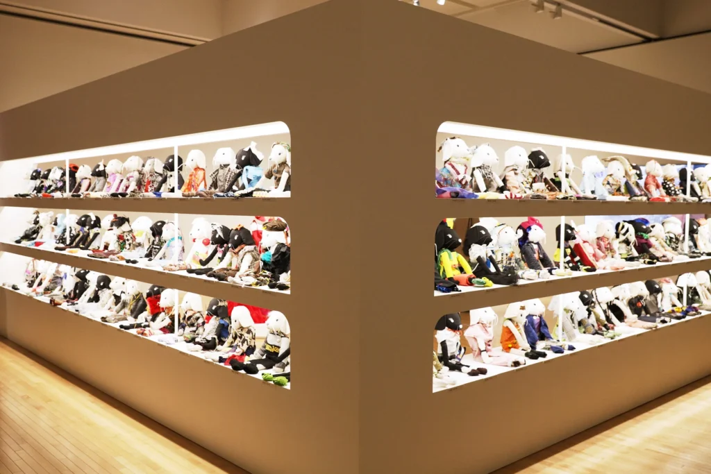 Immersive exhibition installation at the (UN)KNOWN HIROKO KOSHINO retrospective featuring a large sculptural display wall with illuminated recessed shelves filled with rows of miniature mannequin forms dressed in diverse textile compositions, experimental garments, and colorful material studies arranged throughout the contemporary gallery space