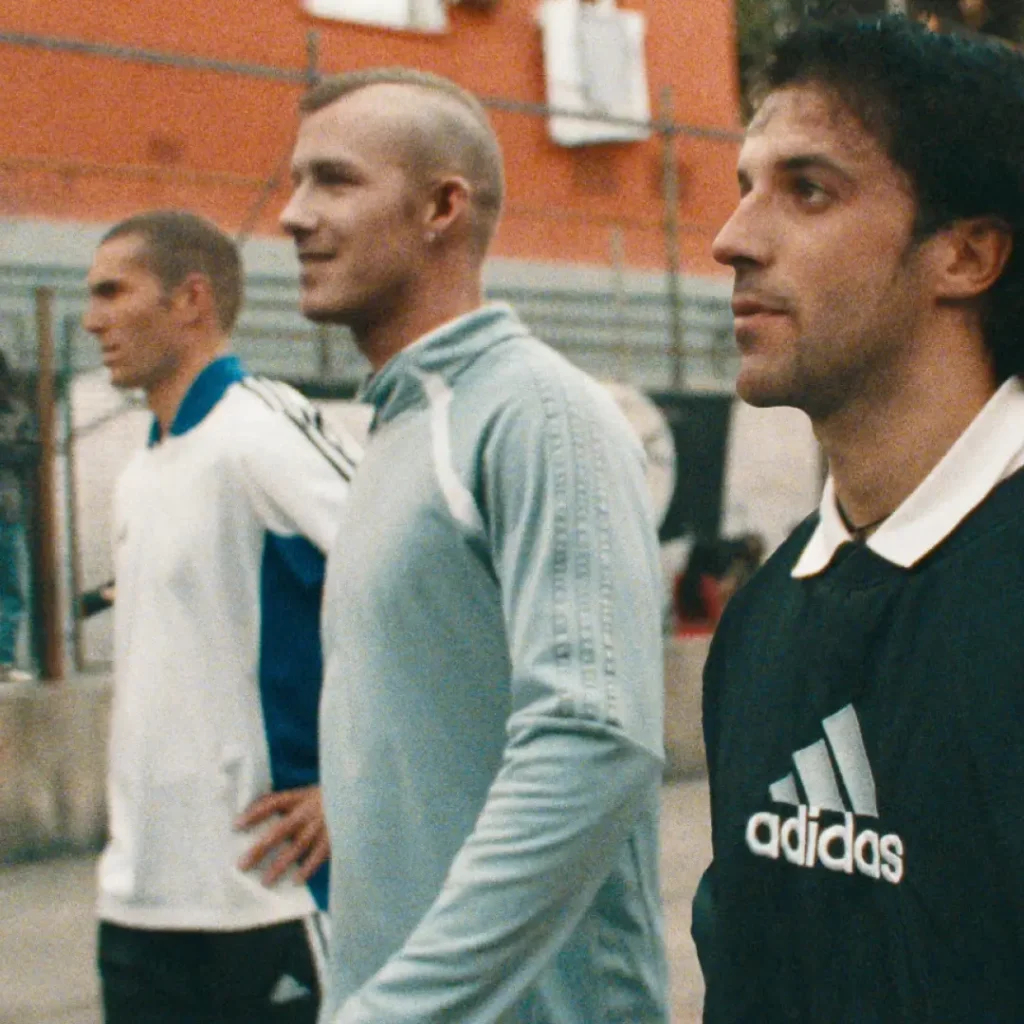 Three men stand side by side on an outdoor football court wearing retro-inspired adidas sportswear in a softly grainy cinematic scene. The central figure wears a pale blue zip-up track top with tonal three-stripe detailing, while the man in the foreground sports a dark adidas football sweatshirt layered over a crisp white collar. Another figure in the background wears a white and blue athletic jersey, reinforcing the archival football aesthetic. Warm muted tones, shallow focus, and urban apartment surroundings create a nostalgic atmosphere reminiscent of classic European street football culture and vintage sports campaigns