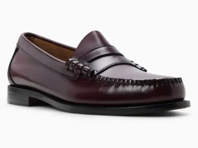 Full side-angle view of a classic penny loafer crafted in glossy oxblood leather, featuring hand-stitched moc toe detailing, a traditional saddle strap, low stacked heel, and sleek black outsole against a light gray studio backdrop