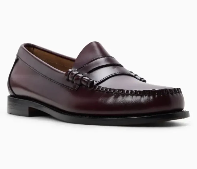 Full side-angle view of a classic penny loafer crafted in glossy oxblood leather, featuring hand-stitched moc toe detailing, a traditional saddle strap, low stacked heel, and sleek black outsole against a light gray studio backdrop