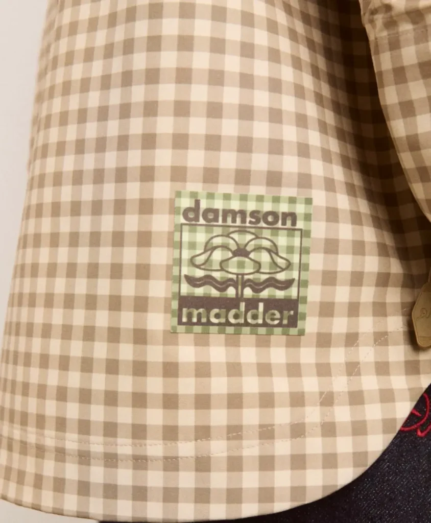 A close-up detail of a beige-and-cream gingham garment featuring a sewn-on square “damson madder” patch in muted green tones. The label carries a stylized floral graphic with retro-inspired typography, reinforcing the British brand’s soft vintage-meets-contemporary aesthetic. Fine stitching curves along the lower hem, while a glimpse of dark indigo denim and a brass zipper pull adds layered texture and tonal contrast to the composition
