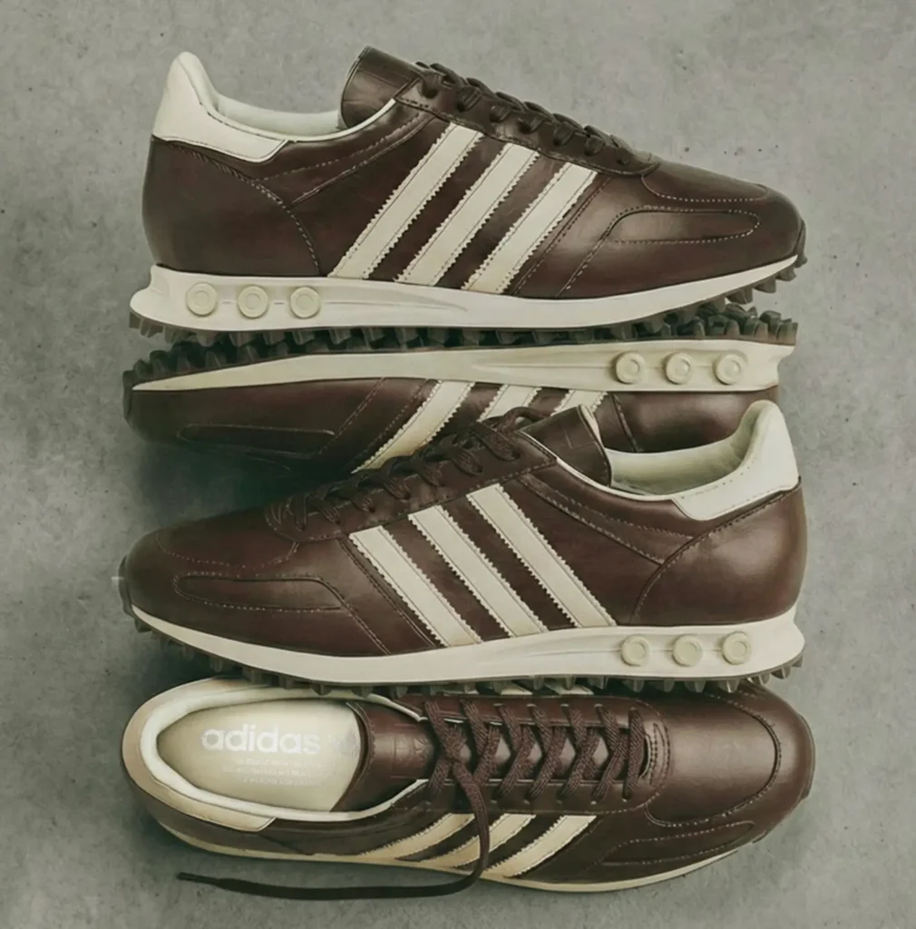 Multiple pairs of the adidas L.A. Trainer “Made in Italy” edition are arranged in a stacked flat-lay composition, showcasing the sneaker’s dark brown leather construction, cream Three Stripes detailing, and signature heel peg cushioning system against a muted industrial backdrop