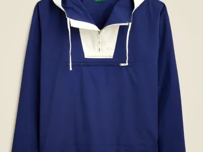 Product image of the J.Crew 1989 Heritage Anorak in a rich navy blue colorway with contrasting white hood and half-zip placket detailing. The lightweight pullover jacket features long white drawstrings, a horizontal zip kangaroo-style pocket across the chest, relaxed sleeves, and adjustable hem ties at the bottom. Displayed against a clean neutral backdrop, the silhouette reflects the archival late-1980s J.Crew aesthetic—functional, minimalist, and rooted in classic American sportswear with a distinctly coastal-prep sensible