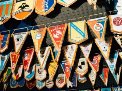 A dense collection of vintage football pennants hangs from layered cords beneath a textured black ceiling, creating a colorful canopy of European club history. Triangular and shield-shaped banners in faded reds, blues, yellows, and greens feature old crests, sponsor marks, and regional team names, all trimmed with gold fringe that adds a nostalgic, trophy-room atmosphere to the scene