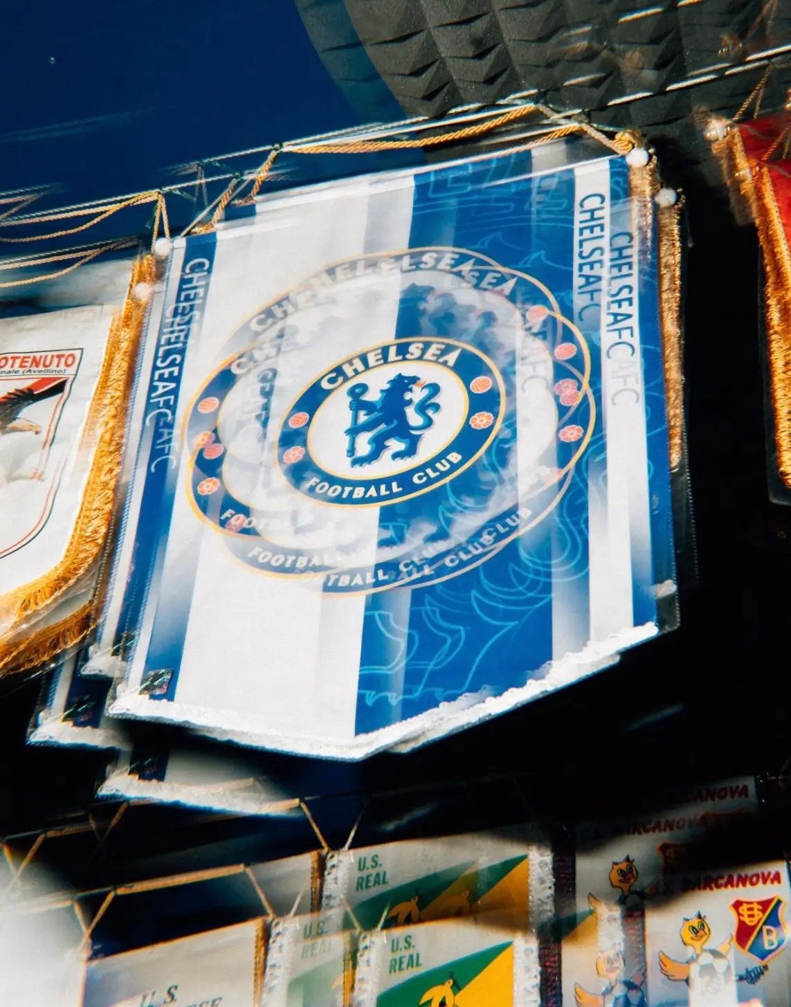 Vintage-style Chelsea FC pennants hang closely together beneath a dark stadium-like ceiling, captured with a blurred, double-exposure effect that gives the blue-and-white crests a layered, dreamlike motion. Gold fringe detailing catches the light along the edges, while smaller retro football banners sit partially visible below, reinforcing the nostalgic supporter-club atmosphere