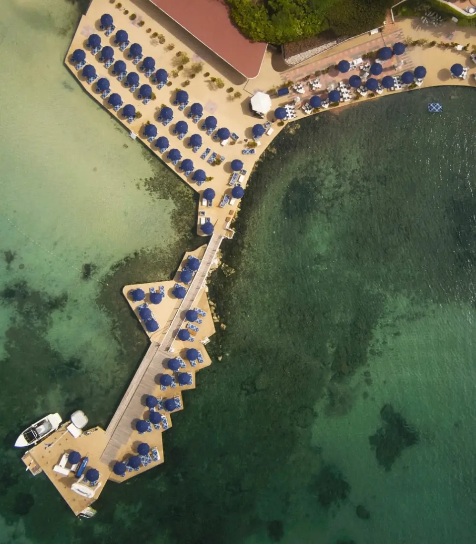 An aerial view of Hôtel Belles Rives reveals Burberry’s Riviera takeover unfolding across the Mediterranean shoreline, where rows of deep blue parasols and check-detailed loungers extend along the hotel’s iconic private jetty and beach club terraces. Surrounded by crystalline turquoise water, the geometric layout emphasizes the immersive scale of the activation, transforming the historic Côte d’Azur destination into a fully branded summer landscape rooted in luxury travel, nautical leisure, and Burberry’s High Summer 2026 aesthetic