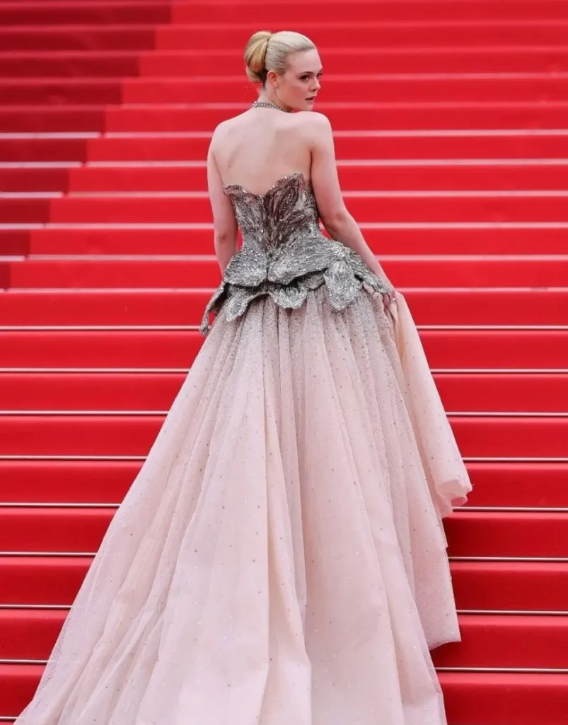 A dramatic couture moment unfolds on the red staircase of the Cannes Film Festival as the subject appears in a voluminous blush-toned gown that balances softness with sculptural intensity. The dress features a sweeping tulle skirt covered in subtle sparkling embellishments, creating a light, ethereal movement against the vivid crimson backdrop. The corseted upper section contrasts sharply with the airy lower half through intricate metallic embroidery that resembles silver petals or molten floral armor. Structured peplum detailing around the hips gives the silhouette an architectural quality, transforming the look from traditional princess couture into something more editorial and avant-garde. Styled with a sleek bun and minimal accessories, the overall presentation allows the gown itself to dominate the frame. The turned-back pose emphasizes the dramatic structure of the bodice while reinforcing the cinematic elegance associated with Cannes red-carpet fashion. The image captures the modern luxury aesthetic perfectly: romantic, theatrical, and engineered for visual impact at scale