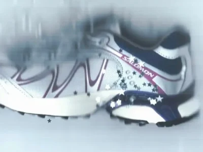 Close-up campaign image of the Salomon x Ray BEAMS sneaker shown in motion against a cool-toned blurred backdrop. The shoe features silver mesh paneling with flowing purple overlays, navy accents, and subtle star embellishments scattered across the frame, emphasizing a futuristic trail-runner aesthetic blended with play Y2K-inspired style