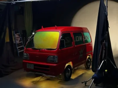 A bright red micro-van sits beneath a suspended studio light rig during what appears to be a campaign or editorial production setup. The compact van features reflective yellow-green windshield tinting, blacked-out wheels, and playful devil horn accents mounted on the roof, reinforcing its eccentric personality. Draped black curtains, exposed lighting equipment, and soft spotlighting create a cinematic behind-the-scenes atmosphere, while subtle branding across the side windows connects the vehicle to contemporary streetwear and technical fashion culture. The image captures the chaotic charm and underground creativity often associated with customized Japanese kei vans and urban automotive storytelling