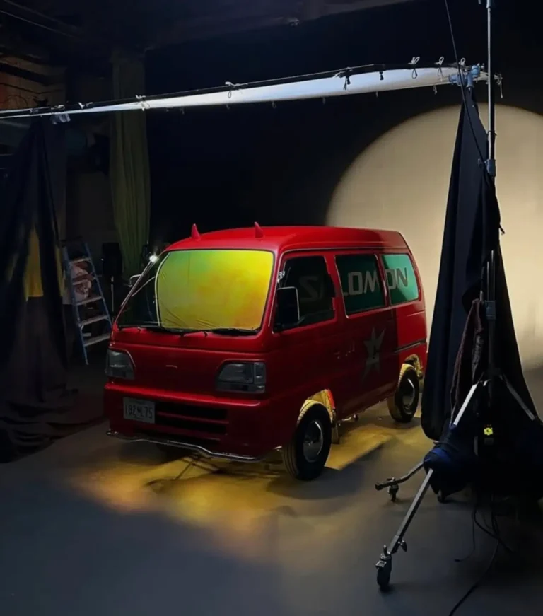 A bright red micro-van sits beneath a suspended studio light rig during what appears to be a campaign or editorial production setup. The compact van features reflective yellow-green windshield tinting, blacked-out wheels, and playful devil horn accents mounted on the roof, reinforcing its eccentric personality. Draped black curtains, exposed lighting equipment, and soft spotlighting create a cinematic behind-the-scenes atmosphere, while subtle branding across the side windows connects the vehicle to contemporary streetwear and technical fashion culture. The image captures the chaotic charm and underground creativity often associated with customized Japanese kei vans and urban automotive storytelling