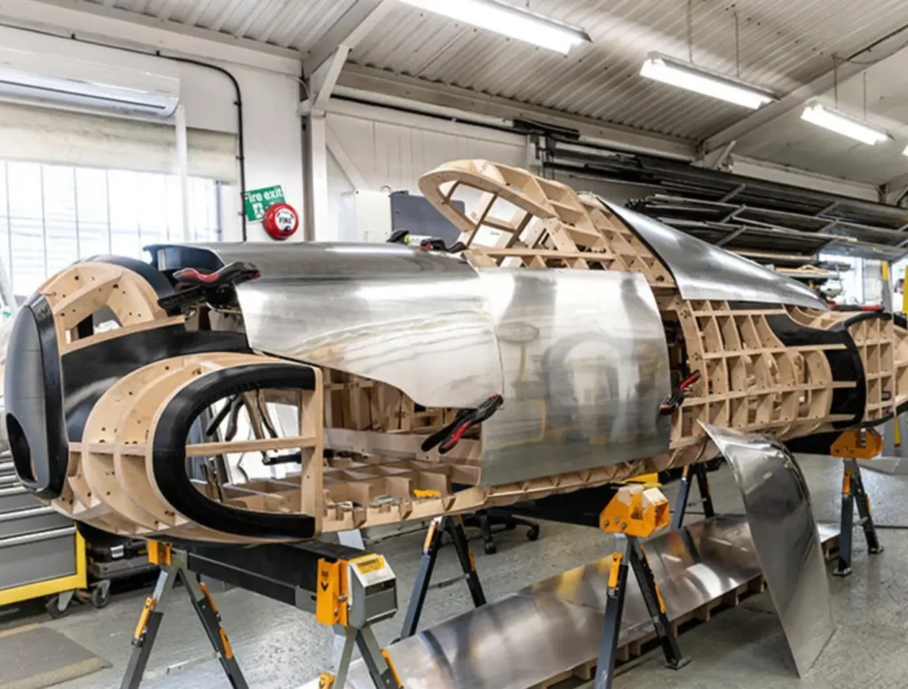 A partially constructed automotive body sits elevated inside an industrial workshop, revealing an intricate wooden skeletal framework beneath unfinished brushed-metal exterior panels. Large circular wheel arches and aerodynamic contours hint at a futuristic or coachbuilt performance car, while clamps, exposed supports, and loose aluminum sheets emphasize the meticulous hand-formed fabrication process underway beneath bright fluorescent lighting