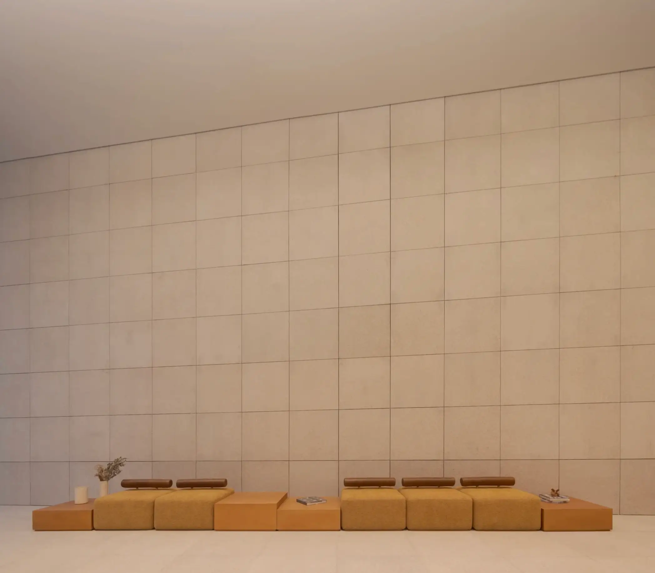 A minimalist interior composition features a long modular floor sofa in muted camel and tan tones positioned against an expansive grid-paneled wall in soft beige. The low-profile seating arrangement, accented by slim leather headrests, books, and sparse decorative objects, emphasizes calm symmetry, restrained luxury, and contemporary architectural minimalism within the oversized monochromatic space