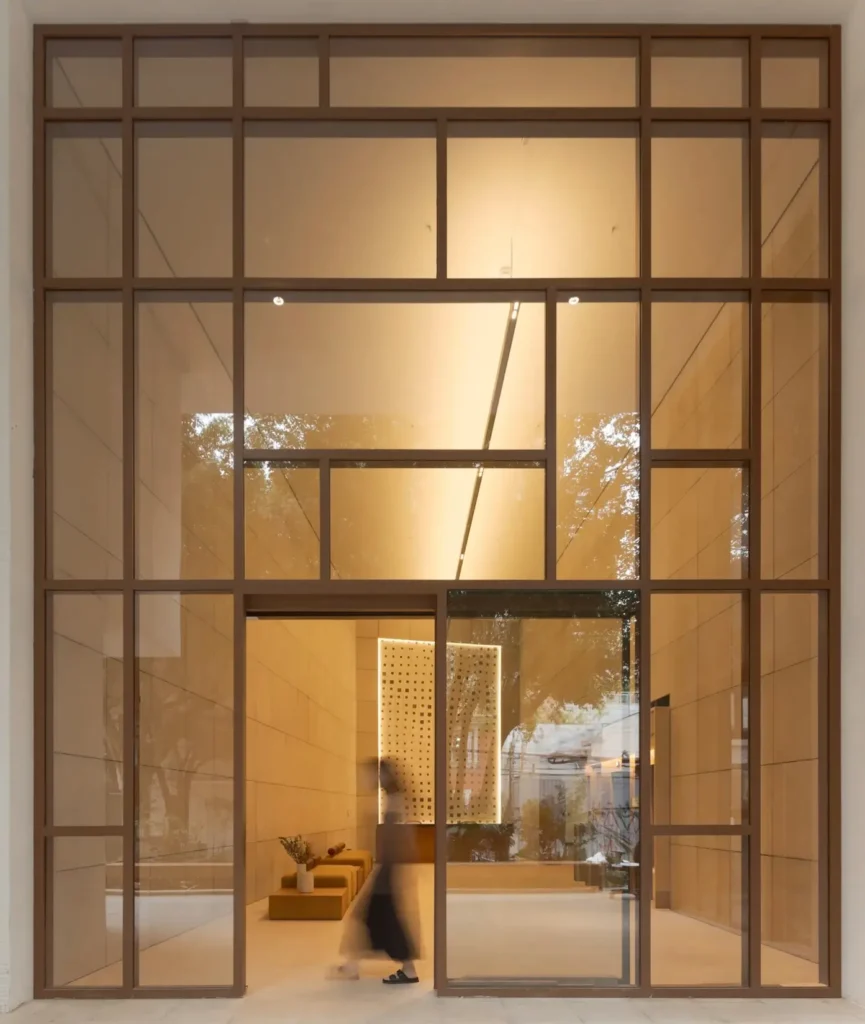 A towering floor-to-ceiling glass façade framed in bronze-toned metal reveals a serene minimalist interior washed in warm ambient lighting. Inside, soft beige wall panels, low modular seating, and a perforated illuminated partition create a refined architectural atmosphere, while the blurred silhouette of a person walking through the entrance adds motion and scale to the calm contemporary space