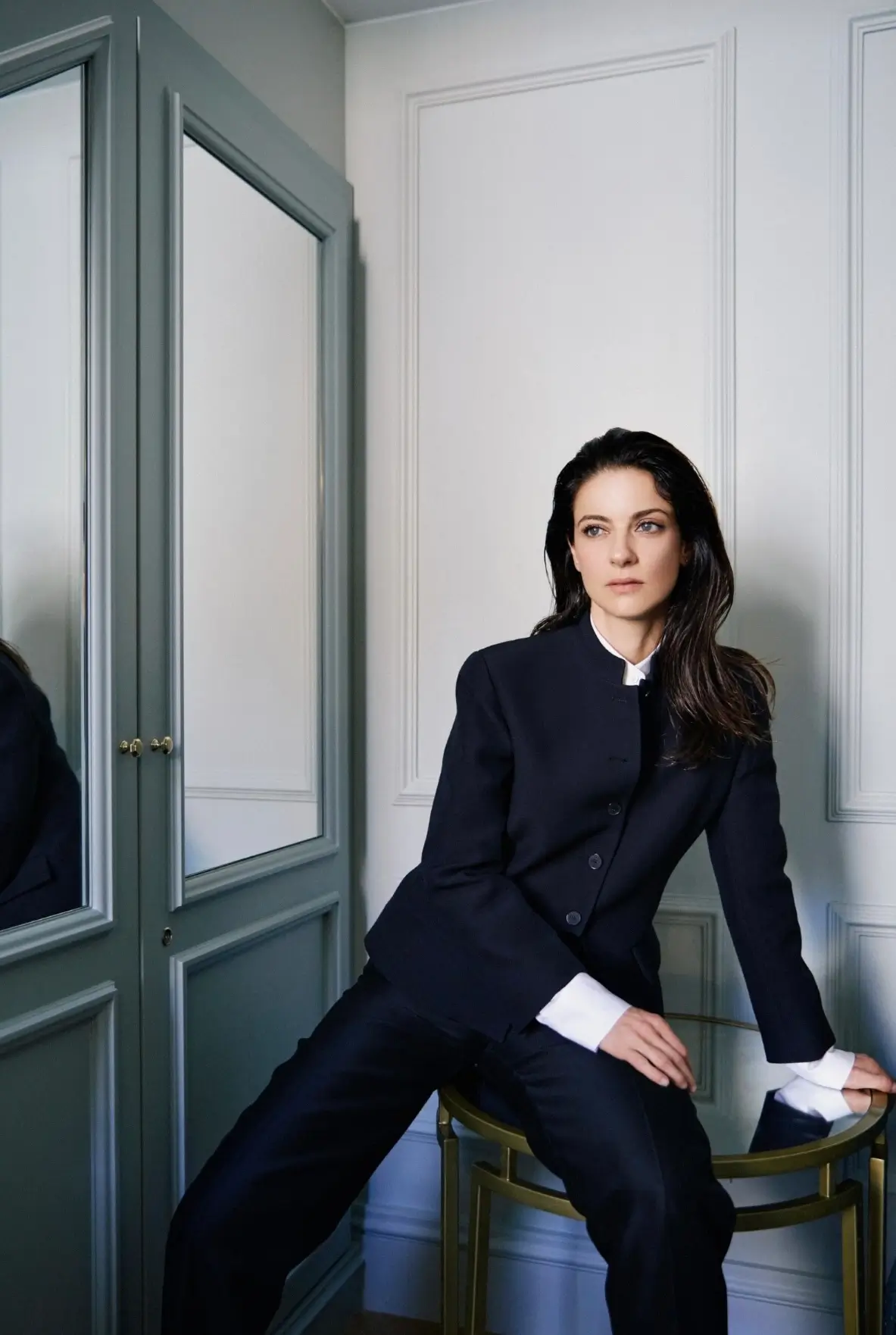 Woman posed in a softly lit Parisian-style interior wearing a sharply tailored dark navy suit with wide-leg trousers and crisp white cuffs, leaning against a mirrored side table with a calm, contemplative expression